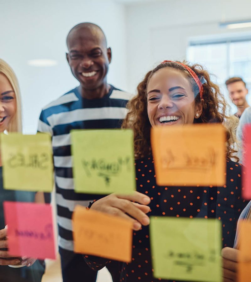 Diverse group of laughing young designers standing in an office brainstorming together with sticky notes on a glass wall